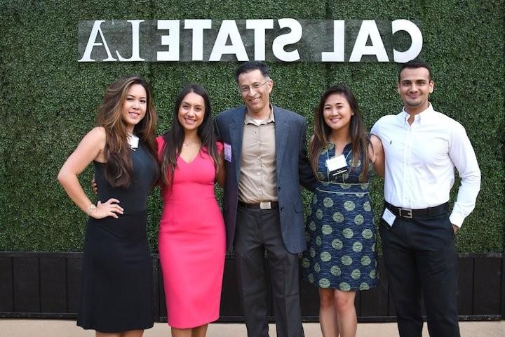 Five smiling alumni posing in front of Cal State LA banner